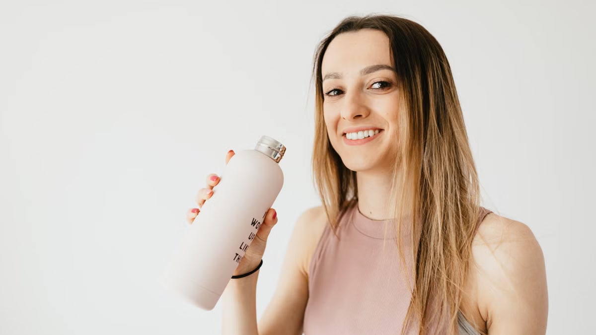 Positive slim woman in pale pink blouse and wristband holding big matte glass bottle with drink while standing in white background and looking at camera